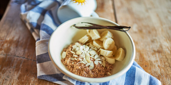 Basenmüsli mit Bananen und Kakao | © SONNENTOR Auf dem Foto sieht man eine Schale mit Basenmüsli. Darauf sind Bananenstücke und Mandelblättchen zu sehen. Auf der Schale liegen Vanilleschoten. | © SONNENTOR